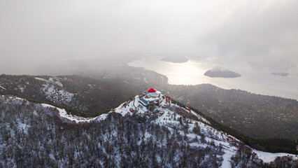 Aerial view of Cerro Otto snowy summit and panoramic lake with clouds and islands in Bariloche, Argentina.