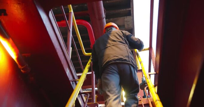 Sailor in helmet climbs steep steel ladder toward bright upper deck. Sun glare reflects from metal wall framing silent determination