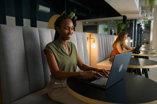Happy young black businesswoman in smart casual using laptop at desk in office lobby
