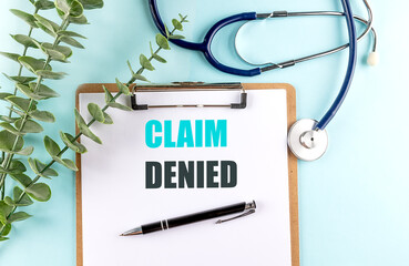 Claim denied message on medical clipboard with stethoscope, pen and eucalyptus on blue background