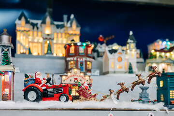 Santa drives a tractor pulling a sleigh through a festive holiday village scene at night