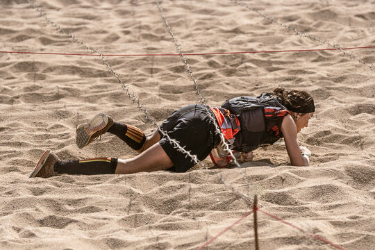 Female athlete crawling on all fours on the sand under barbed wire on an outdoor obstacle course at the beach.