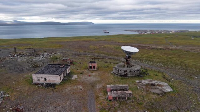 The camera performs a slow orbit around the parabolic antenna of the Orbita station in Chukotka, exposing mechanisms, stairway and circular base while the bay gleams beyond The scene feels remote and