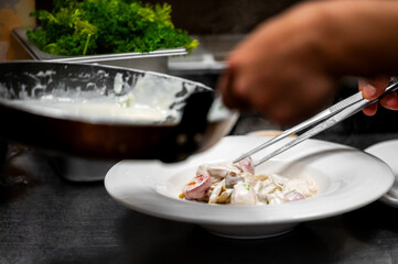 Chef in a professional kitchen uses metal tongs to plate a serving of creamy seafood or pasta with red onions and mushrooms onto a white plate. Shallow depth of field, action shot.