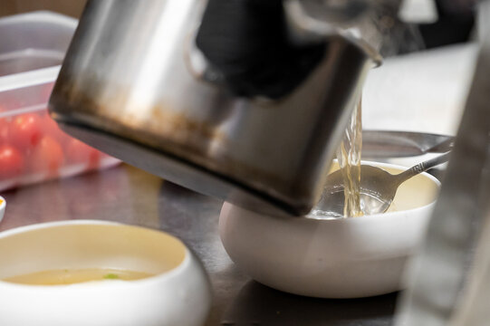 Professional chef pouring hot broth or stock from a stainless steel pot into a small white serving bowl with a silver ladle in a commercial kitchen setting