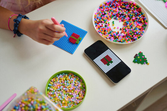 perler bead craft activity with colorful beads, smartphone app guidance, and creative design on tabletop. woman's hand placing beads on blue pegboard for vibrant art project