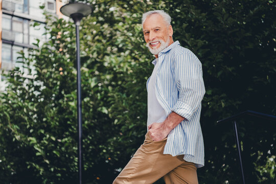 Senior man in casual summer outfit walks in a city park beside apartment buildings under daylight sun