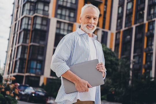 Elderly man outdoors in city holding laptop smiling in front of modern apartment buildings urban summer day