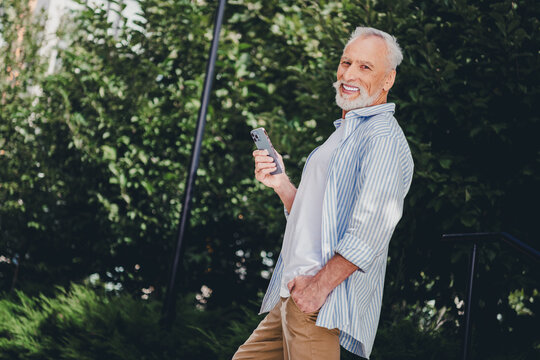 Happy senior man outdoors with smartphone in casual striped shirt enjoying sunny park setting conveying modern lifestyle and real estate interest for stock imagery