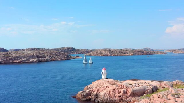 Aerial drone fast flight over ocean and sailboats passing lighthouse on a rocky coast. Two sail yachts approaching lighthouse aon sunny summer day. Sweden west coast between islands