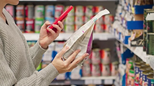 Woman scanning dry pet food using smartphone in pet store. Modern customer checking product information and ingredients using mobile app, AI assistant. Smart shopping Conscious choice Digital retail