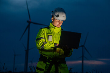 A man in a yellow jacket is looking at a laptop. He is wearing a hard hat. Engineers working at night using a laptop for wind turbine maintenance and monitoring.