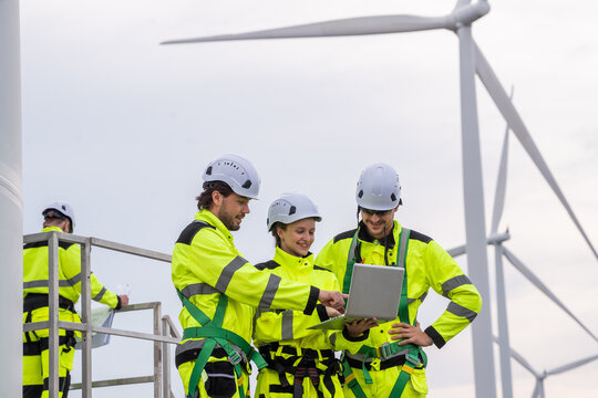 Three people wearing yellow safety gear are looking at a laptop. One of them is wearing a hard hat. Professional workers collaborating on renewable energy project, focusing on safety and maintenance.