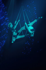 Female aerial duo on chains under teal spotlight—performing aerial tricks with strength, elegance and control; vertical frame. 