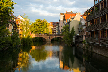 Golden hour over the Pegnitz in Nurnberg, Germany, glows warmly. The historic Maxbr&uuml;cke spans the river like a calm anchor between leafy banks. Reflected facades amplify slow evening magic.