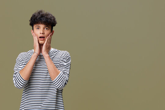surprised young man in striped shirt with hands on cheeks, dark curly hair against plain olive background, casual expression, emotional reaction