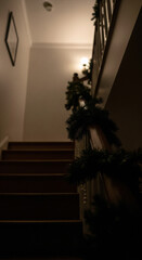 Atmospheric low-angle view of a home staircase decorated with a Christmas garland. A moody, dark scene leading to a warm light, evoking the quiet anticipation and magic of Christmas Eve.