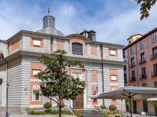 Fototapeta premium The historic Iglesia de San Nicolás de los Servitas in central Madrid, with its brick-and-stone facade and dome, sits next to a square with a small tree and an outdoor café awning