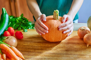 A woman holds a ripe pumpkin with her hands, ripe vegetables on a wooden table. Autumn background with space for copy, ripe vegetables, tomatoes, onions, zucchini, herbs, space for text and