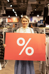 The girl is holding a big red banner with a percent sign and smiling. The girl in the clothing store shows a big discount, sale. Black Friday
