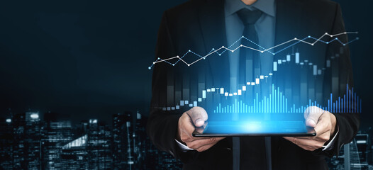 A businessman in a suit holds a tablet displaying vibrant financial graphs and data visualizations against a night city backdrop, symbolizing economic growth and market analysis. Scalp