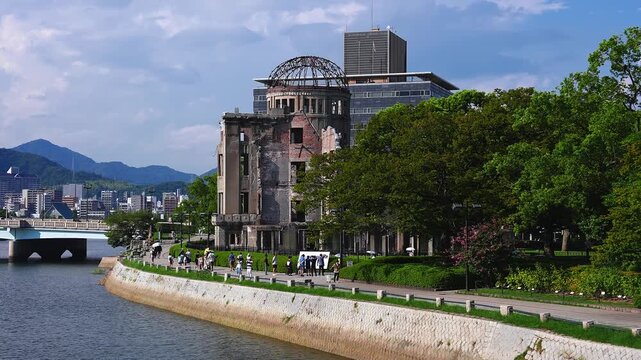 Daytime view of the A Bomb Dome on the Motoyasu River, visitors stroll with umbrellas, blue bridge and city buildings behind, park trees and low mountains in Hiroshima, Japan.