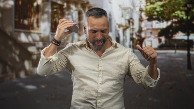 Man middle aged with grey beard holding phone to ear on a city street while dancing and gesturing with raised fist and open hand; playful.