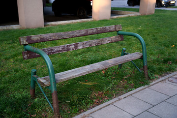 weathered wooden bench with green metal frame on vibrant lawn, next to urban sidewalk, illuminated...