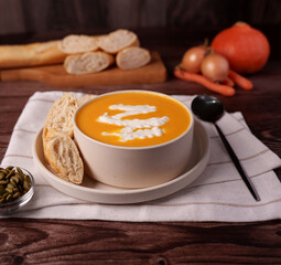 creamy pumpkin soup with whipped cream drizzle, accompanied by freshly baked bread slices and rustic vegetables, presented on a wooden table setting for cozy autumn meal, arrangement, artisanal