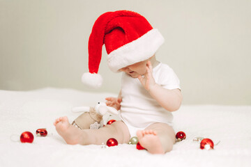 Baby sitting on the bed wearing a Santa hat, surrounded by festive bokeh and Christmas ornaments. The child is exploring red and gold decorations with curiosity and joy.