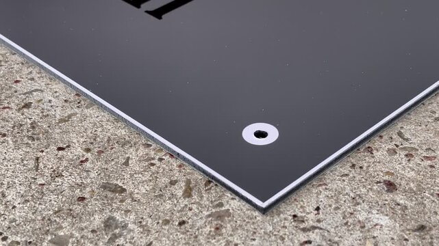 Close-up of modern black composite aluminum sign panel corner with precision-drilled mounting hole on concrete surface &mdash; minimalist industrial design detail symbolizing craftsmanship