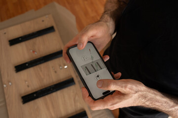 Man checking furniture assembly instructions on smartphone, soft light. Male hands holding phone with illustrated guide above wooden board with metal rails and screws