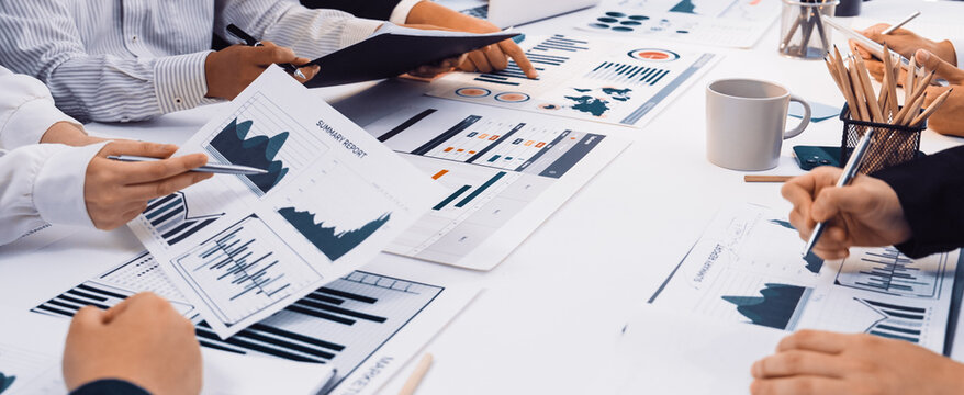 A diverse business team engages in a detailed discussion over graphs and charts displayed on paper and digital devices, fostering collaboration and strategy development. SACTR - Powered by Adobe