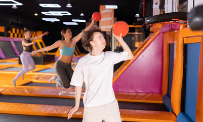 Positive young man jumping on trampoline to throw ball through ring in modern hall