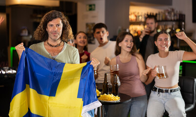 Company of excited sports fans waving flag of Sweden and supporting national team with beer in bar