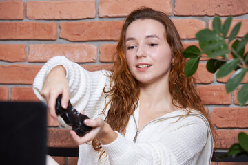A young beautiful girl sits on the sofa holding a gamepad in her hands and playing a computer game. The player rejoices in victory