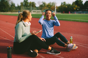 Two diverse female friends and athletes, one Asian and one Caucasian, take a joyful break on a running track, listening to music and chatting after a workout.
