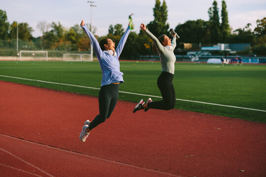 Energetic diverse female friends and athletes, one Asian and one Caucasian, jump for joy and celebrate success on a running track after completing a workout