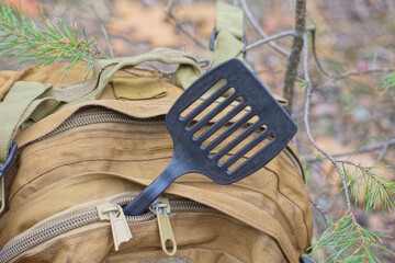 one black plastic large slotted spoon lies in the pocket of a brown backpack on a summer street