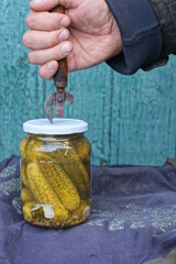 A hand with an old rusty can opener opens a white metal lid on a glass jar with green pickled cucumbers