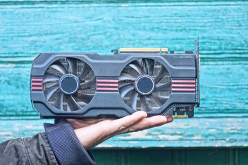 A hand holds one large black video card with two fans against a green wall on a summer street