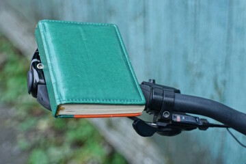 one small closed green notepad with leather cover lies on a black bicycle handlebar on a summer street