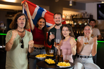 Group of friends, excited supporters cheering for favorite team with flag of Norway while watching match on TV in bar