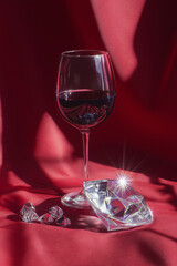 Glass of red wine and large clear diamond with smaller crystals reflecting light on a elegant red silk fabric background