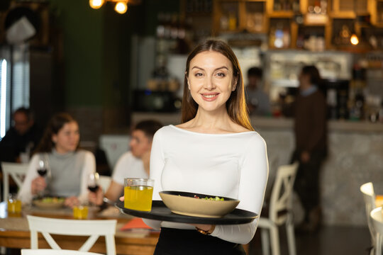 Girl waiter with tray walks between rows of tables. Employee of public catering cafe carries order to client, ready to serve guest of cafe establishment.
