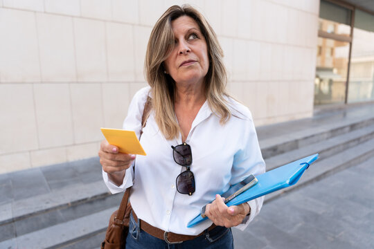 A middle-aged woman on the street wearing a shirt and holding a note, looking around for a financial building