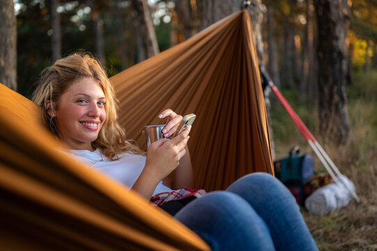 Woman relaxing in hammock, smiling, using phone while camping