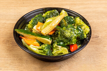 A black takeout bowl of Chinese stir fry vegetables including baby corn, broccoli, zucchini and peppers