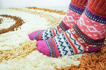 Female feet in warm socks with Nordic ornament resting on soft carpet surface. Reflects sustainable fashion, slow craft movement, natural materials, winter hygge mood, and calm domestic atmosphere.