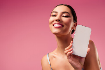 Gorgeous young woman holding smartphone in studio against pink background showcasing style and modern technology
